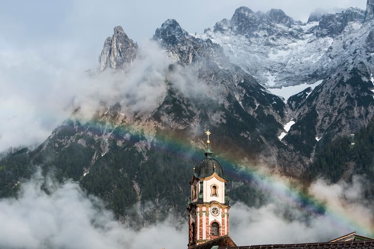 Unbeständiges Wetter erwartet uns am ersten Maiwochenende in den Bergen. Foto: Regenbogen über dem Kirchturm von Mittenwald im bayerischen Karwendel