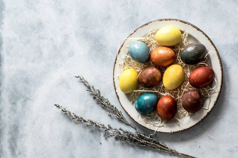 grauer Hintergrund mit weißem Teller, bunt gefärbte Eiern und einem getrocknetem Strauß Rosmarin